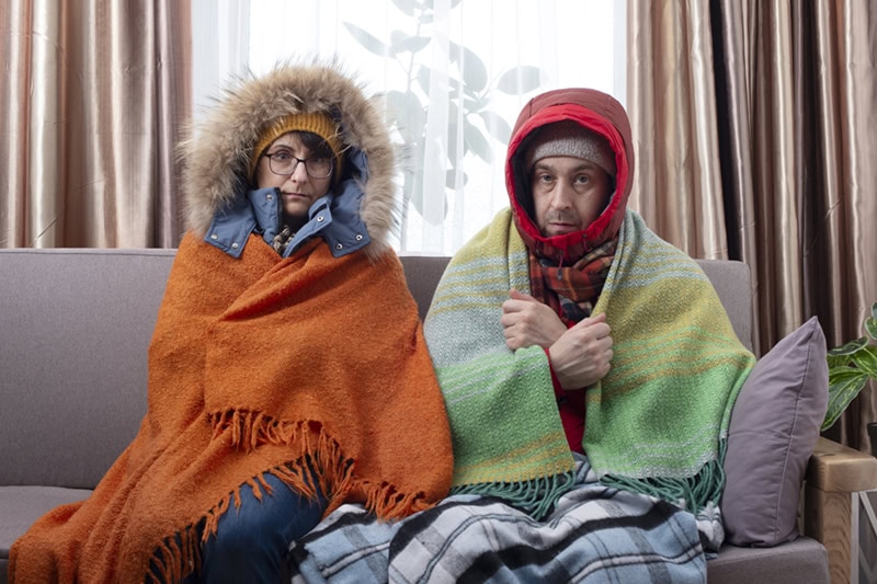 Couple bunded up and sitting on their couch; cold because of bad cold spots in their home.