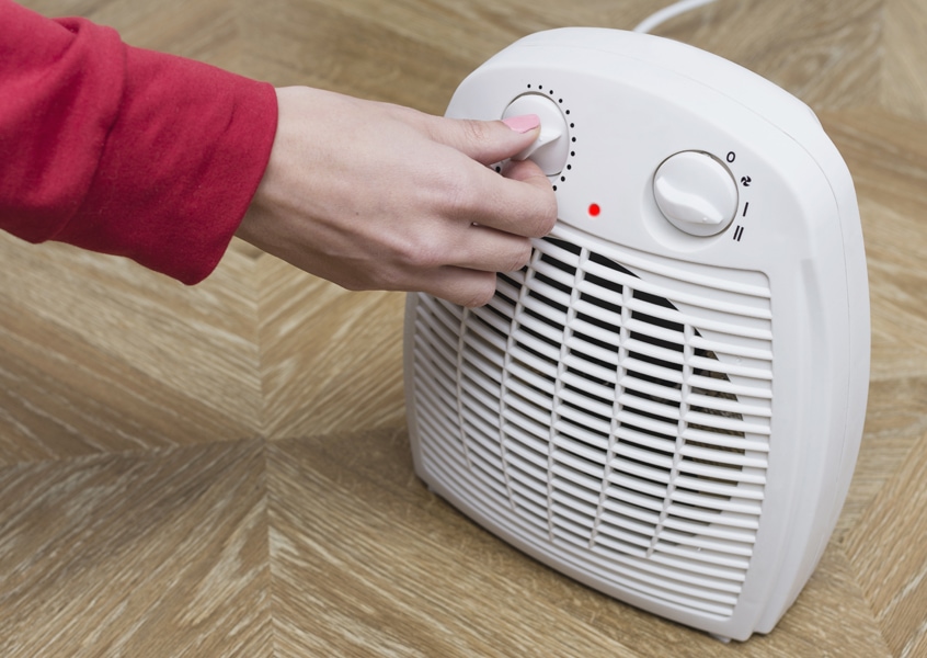 Woman warms her frozen hands near an electric fan heater at home. How can I save money on my heating bill this winter?
